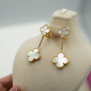 May include: A pair of gold-coloured clover-shaped earrings with white mother-of-pearl centres. One earring hangs from a chain, while the other is a stud. The earrings are displayed on a beige mannequin head.
