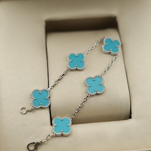 Puede incluir: Dos pulseras plateadas con abalorios turquesas en forma de tr&eacute;bol. Las pulseras se exhiben dentro de una caja de joyer&iacute;a beige, mostrando el dise&ntilde;o delicado y el contraste de color.