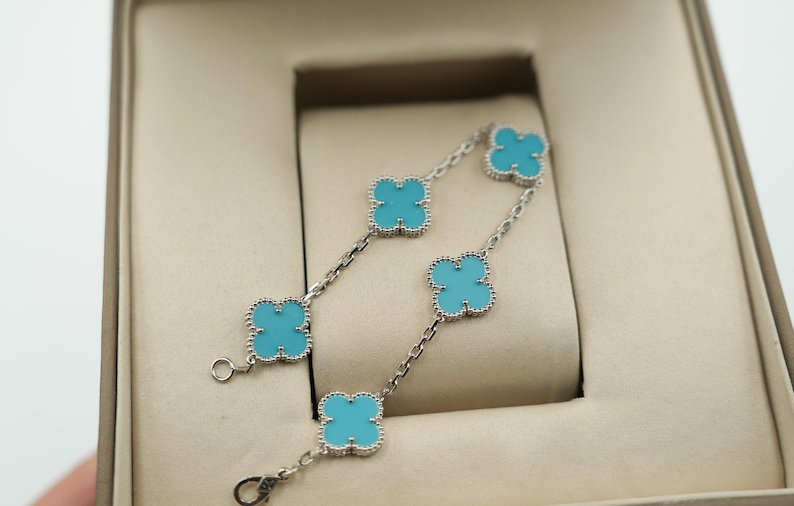 Puede incluir: Dos pulseras plateadas con abalorios turquesas en forma de tr&eacute;bol. Las pulseras se exhiben dentro de una caja de joyer&iacute;a beige, mostrando el dise&ntilde;o delicado y el contraste de color.