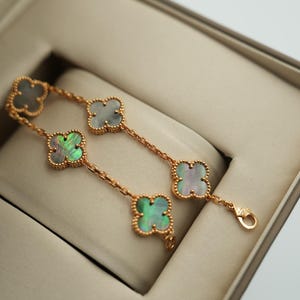 K&ouml;nnte beinhalten: Ein goldfarbenes Armband mit vierbl&auml;ttrigen Kleeblattmotiven mit schillernden Perlmuttzentren. Das Armband wird in einer beigen Schmuckschatulle pr&auml;sentiert, die die zarte Kette und den Verschluss hervorhebt.