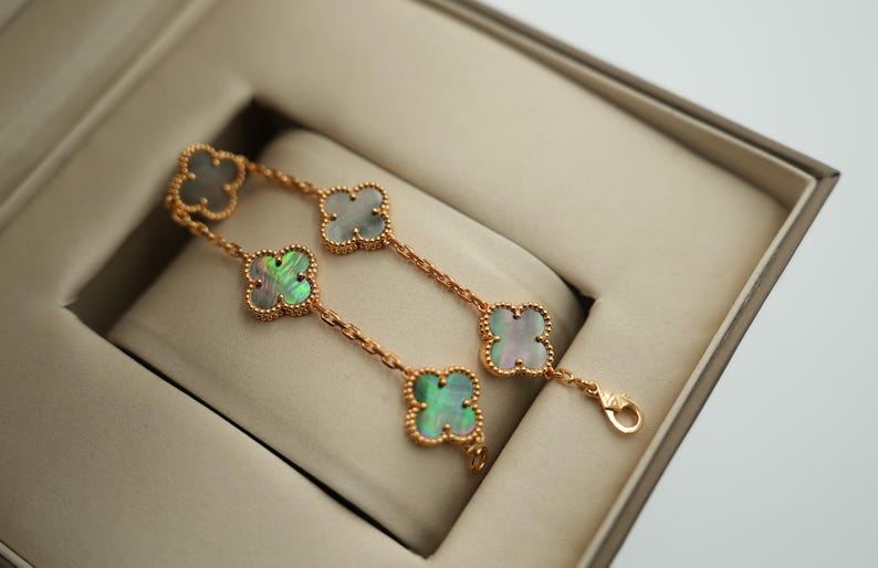 K&ouml;nnte beinhalten: Ein goldfarbenes Armband mit vierbl&auml;ttrigen Kleeblattmotiven mit schillernden Perlmuttzentren. Das Armband wird in einer beigen Schmuckschatulle pr&auml;sentiert, die die zarte Kette und den Verschluss hervorhebt.
