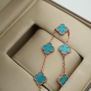 Puede incluir: Pendientes de oro rosa con dise&ntilde;os de tr&eacute;boles de cuatro hojas y centros turquesa. Se exhiben en un joyero beige, mostrando su delicada cadena y detalles intrincados. Un accesorio elegante.