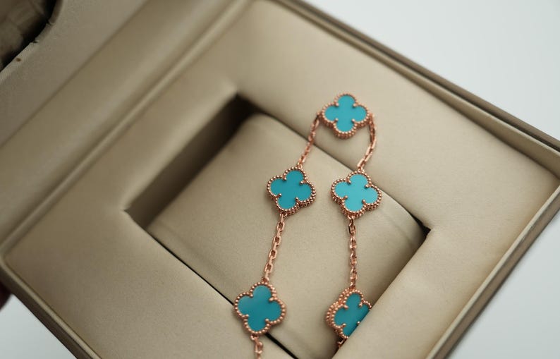 Puede incluir: Pendientes de oro rosa con dise&ntilde;os de tr&eacute;boles de cuatro hojas y centros turquesa. Se exhiben en un joyero beige, mostrando su delicada cadena y detalles intrincados. Un accesorio elegante.
