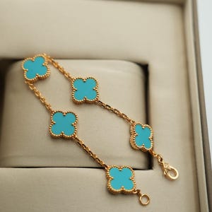 Puede incluir: Pulsera dorada con abalorios turquesas en forma de tr&eacute;bol. La pulsera se muestra en una caja de tonos neutros, resaltando el contraste de colores. El dise&ntilde;o de tr&eacute;bol es un motivo cl&aacute;sico de joyer&iacute;a.