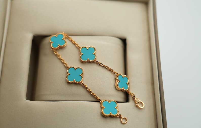 Puede incluir: Pulsera dorada con abalorios turquesas en forma de tr&eacute;bol. La pulsera se muestra en una caja de tonos neutros, resaltando el contraste de colores. El dise&ntilde;o de tr&eacute;bol es un motivo cl&aacute;sico de joyer&iacute;a.