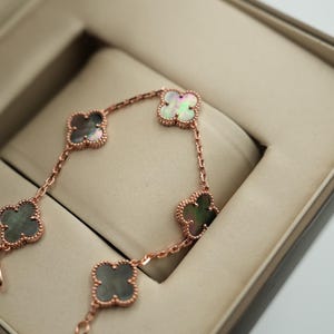 K&ouml;nnte beinhalten: Ein ros&eacute;goldenes Armband mit f&uuml;nf Kleeblatt-Anh&auml;ngern, jeweils mit einem schillernden Perlmutt-Zentrum. Das Armband wird in einer offenen, beigefarbenen Schmuckschatulle mit gepolstertem Innenraum pr&auml;sentiert. Das Design hebt die filigranen Details hervor.