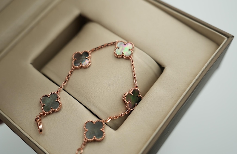 K&ouml;nnte beinhalten: Ein ros&eacute;goldenes Armband mit f&uuml;nf Kleeblatt-Anh&auml;ngern, jeweils mit einem schillernden Perlmutt-Zentrum. Das Armband wird in einer offenen, beigefarbenen Schmuckschatulle mit gepolstertem Innenraum pr&auml;sentiert. Das Design hebt die filigranen Details hervor.