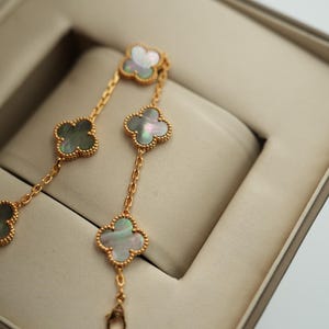 K&ouml;nnte beinhalten: Ein goldfarbenes Armband mit vierbl&auml;ttrigen Kleeblatt-Anh&auml;ngern. Jeder Anh&auml;nger hat einen Perlmutt-Mittelteil und einen goldfarbenen, perlenbesetzten Rand. Das Armband wird in einer beigen Schmuckschatulle pr&auml;sentiert.