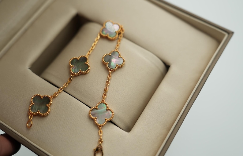 K&ouml;nnte beinhalten: Ein goldfarbenes Armband mit vierbl&auml;ttrigen Kleeblatt-Anh&auml;ngern. Jeder Anh&auml;nger hat einen Perlmutt-Mittelteil und einen goldfarbenen, perlenbesetzten Rand. Das Armband wird in einer beigen Schmuckschatulle pr&auml;sentiert.