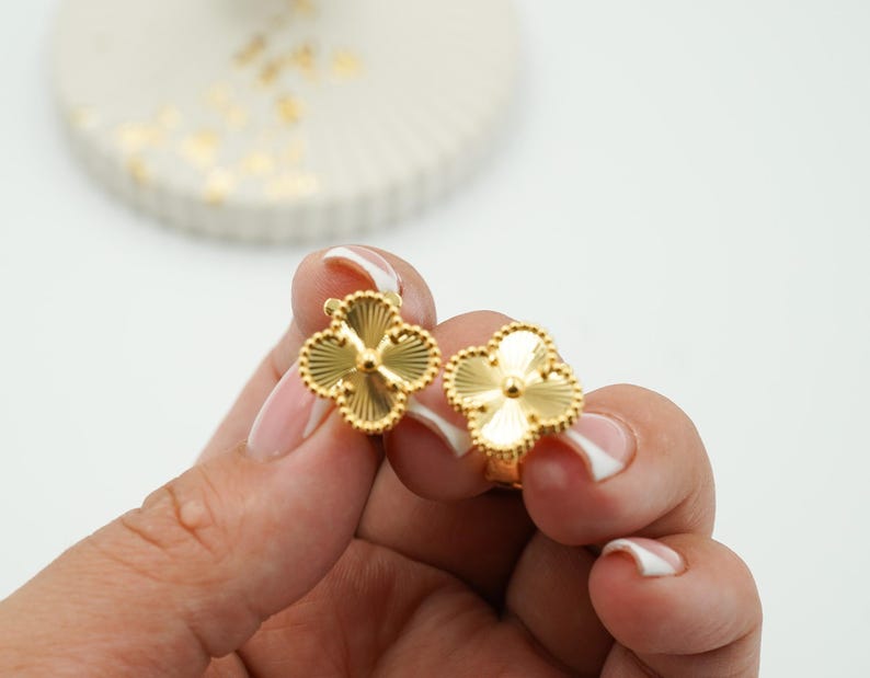 K&ouml;nnte beinhalten: Ein Paar goldfarbene Kleeblatt-Ohrringe. Jeder Ohrring hat vier Bl&uuml;tenbl&auml;tter mit einem strukturierten, geriffelten Design. Die Ohrringe werden vor einem wei&szlig;en Hintergrund gehalten.