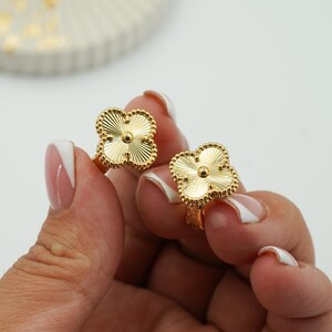 K&ouml;nnte beinhalten: Zwei goldfarbene Ringe in Kleeblattform. Jeder Ring hat ein vierbl&auml;ttriges Kleeblatt-Design mit einer strukturierten, geriffelten Oberfl&auml;che. Die Ringe werden vor einem wei&szlig;en Hintergrund gehalten, um ihre komplizierten Details und ihren Glanz zu zeigen.