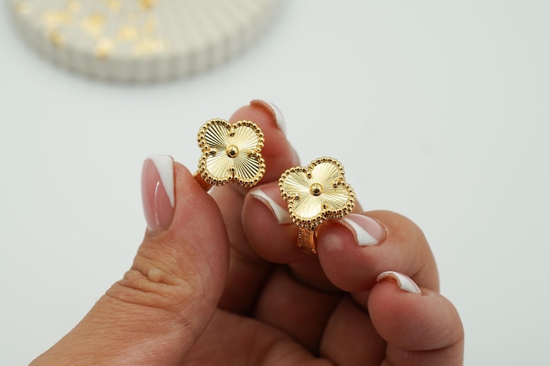 K&ouml;nnte beinhalten: Zwei goldfarbene Ringe in Kleeblattform. Jeder Ring hat ein vierbl&auml;ttriges Kleeblatt-Design mit einer strukturierten, geriffelten Oberfl&auml;che. Die Ringe werden vor einem wei&szlig;en Hintergrund gehalten, um ihre komplizierten Details und ihren Glanz zu zeigen.