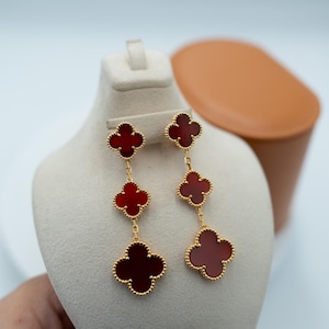 Op de afbeelding: Goudkleurige oorhangers met klaverbladdesigns, elk met een dieprood midden. De oorbellen worden gepresenteerd op een witte buste, waarbij het gedetailleerde ontwerp en de warme rode tint van de klaverhangers worden benadrukt.