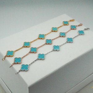 Puede incluir: Cuatro pulseras con piedras turquesas en forma de tr&eacute;bol. Las pulseras son doradas, oro rosa y plateadas. Las pulseras se exhiben sobre una superficie blanca.