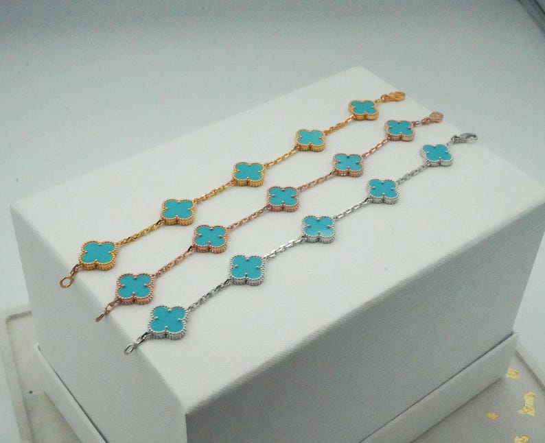 Puede incluir: Cuatro pulseras con piedras turquesas en forma de tr&eacute;bol. Las pulseras son doradas, oro rosa y plateadas. Las pulseras se exhiben sobre una superficie blanca.