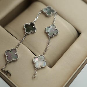K&ouml;nnte beinhalten: Ein silbernes Armband mit Kleeblatt-Anh&auml;ngern mit schillerndem Perlmutt und einem Perlenrand. Das Armband wird in einer beigen Schmuckschatulle pr&auml;sentiert, die sein zartes Design und seine Handwerkskunst hervorhebt.