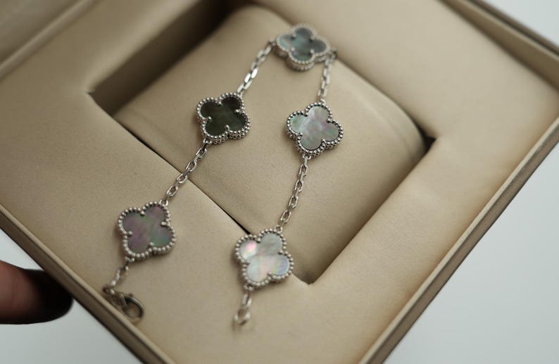 K&ouml;nnte beinhalten: Ein silbernes Armband mit Kleeblatt-Anh&auml;ngern mit schillerndem Perlmutt und einem Perlenrand. Das Armband wird in einer beigen Schmuckschatulle pr&auml;sentiert, die sein zartes Design und seine Handwerkskunst hervorhebt.
