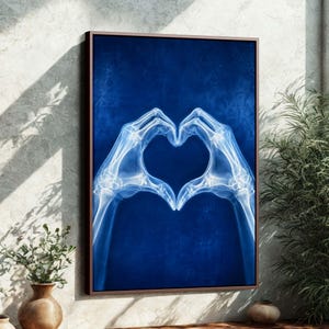 May include: Framed artwork featuring an X-ray image of hands forming a heart shape. The image is in shades of blue and white, with a dark brown frame. The artwork is displayed on a textured wall.