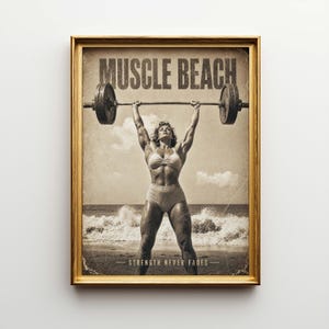 Op de afbeelding: Een ingelijste vintage print in sepia tinten. De afbeelding toont een gespierde persoon die een halter optilt op een strand. De woorden "MUSCLE BEACH" staan bovenaan en "STRENGTH NEVER FADES" onderaan.