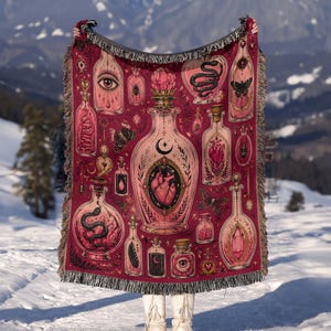 May include: A woven throw blanket featuring a deep red and pink color palette with a repeating pattern of ornate glass bottles and jars. The illustrations include anatomical hearts, snakes, moths, eyes, crystals, and crescent moon symbols.