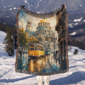 May include: A woven throw blanket featuring a detailed illustration of a yellow tram traveling on tracks in front of a large cathedral with a golden dome. The scene includes city buildings, overhead power lines, and a wet street reflecting the light.