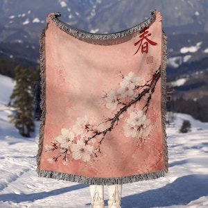 May include: A woven throw blanket featuring a soft pink watercolor background with a dark brown cherry blossom branch extending diagonally. The blanket includes white floral blossoms, a red Chinese character in the top right corner, and a fringed edge.