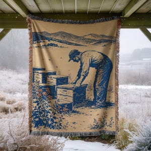 May include: A woven throw blanket featuring a tan background with a blue illustration of a person tending to stacked wooden beehives in a field. The design includes mountains in the background, floral accents, and a fringed edge on all sides.