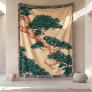 May include: A woven throw blanket featuring a large, detailed illustration of a pine tree with a textured, light tan background and deep green foliage. The tree has a prominent, winding brown trunk. The blanket has fringed edges on all sides.