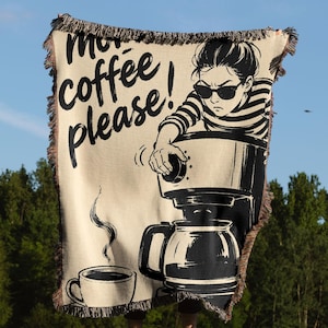 May include: A woven throw blanket featuring a cream background with black graphic illustrations. The design depicts a person wearing sunglasses operating a coffee maker, a steaming cup of coffee, and the text "more coffee please!" with fringed edges.