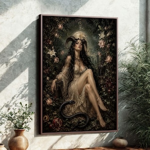 Op de afbeelding: Ingelijste kunstdruk met een vrouw met hoorns, lang donker haar en een vloeiende jurk, zittend te midden van bloemen en bladeren. Een zwarte slang is aan haar voeten opgerold. Het kunstwerk heeft een donkere, gotische esthetiek.