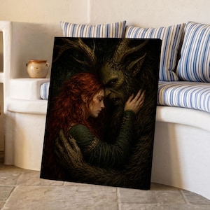 May include: A framed wall art print depicting a person with long red hair wearing a green garment embracing a dark, horned, fur-covered creature. The artwork features deep shadows and earthy tones, encased in a light-colored wooden frame.