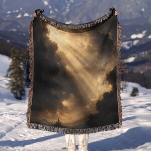 May include: A rectangular woven throw blanket featuring a dark, dramatic sky with golden light rays breaking through heavy clouds above a silhouette of a castle. The blanket has a fringed edge and a textured, tapestry-style weave throughout.