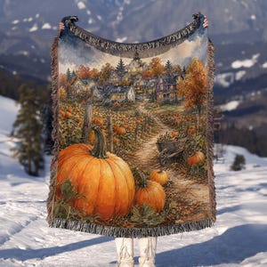 May include: A rectangular woven throw blanket featuring a detailed autumn scene with large orange pumpkins in the foreground, a dirt path leading to a small village with houses, and trees with fall foliage, finished with a fringed edge on all sides.