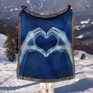 May include: A rectangular woven throw blanket featuring a deep blue background with a central white X-ray graphic of two skeletal hands forming a heart shape. The blanket has a decorative fringed edge along the top and bottom borders.