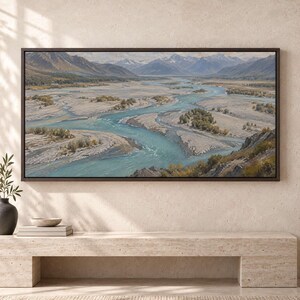 May include: A rectangular landscape painting in a dark brown frame depicting a wide, winding turquoise river flowing through a vast, rocky valley floor, surrounded by distant snow-capped mountains under a clear, bright sky.