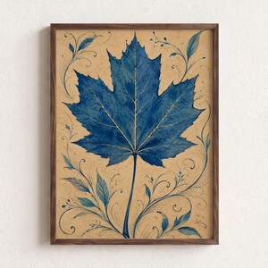 May include: A framed wall art print featuring a large, detailed blue maple leaf centred on a textured tan background. The leaf is surrounded by delicate, swirling blue vine and leaf illustrations that extend towards the corners of the frame.