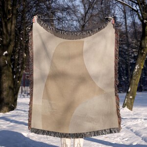 May include: A woven throw blanket featuring a large, abstract beige shape centered on a light gray background. The blanket has a textured appearance with dark, multi-colored fringe along all four edges, creating a distinct border around the design.