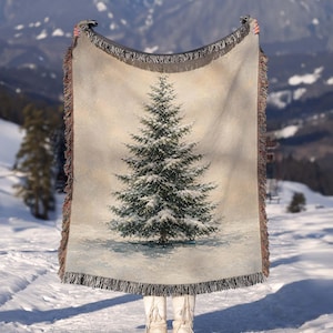 May include: A woven throw blanket featuring a detailed illustration of a snow-covered evergreen tree centered against a soft, neutral background. The rectangular blanket has decorative fringed edges along the sides and bottom, with a textured finish.