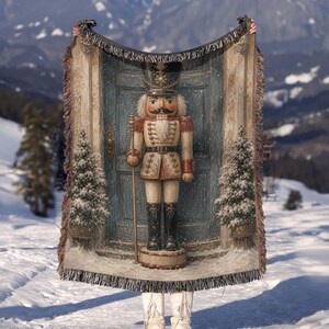 May include: A woven throw blanket featuring a detailed illustration of a nutcracker standing before a blue door flanked by two snow-covered evergreen trees. The design includes a tall hat, a gold staff, and a white uniform with gold accents.