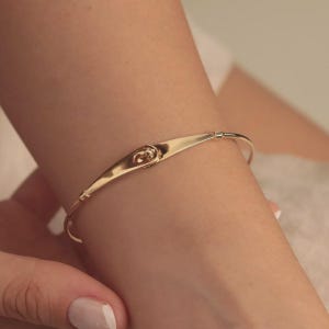 May include: A gold-coloured bangle bracelet. The bracelet features a curved, elongated centrepiece with a decorative element. The thin, circular band is connected to the centrepiece. The bracelet is worn on a wrist.