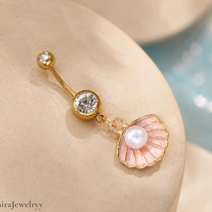 Puede incluir: Anillo para el ombligo dorado con un diseño de barra curva. Tiene una pequeña gema transparente en la parte superior, una gema más grande en el centro y un dije de concha rosa con una perla. Sobre un fondo neutro.