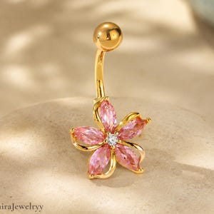 Könnte beinhalten: Ein goldfarbener Bauchnabelpiercing mit einem Blumendesign. Die Blume hat rosa Blütenblätter und einen kleinen, klaren Edelstein in der Mitte. Der Ring ist gebogen und hat oben eine goldene Kugel. Der Markenname "SolmiraJewelryy" steht unten.