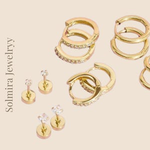 Puede incluir: Conjunto de joyas doradas sobre un fondo neutro. La imagen presenta múltiples pares de pendientes de aro, algunos adornados con pequeños cristales, y varios pares de pendientes de botón con gemas transparentes. El texto "Solmira Jewelryy" es visible.