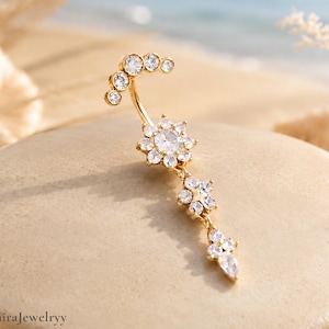 Può includere: Un piercing per l'ombelico color oro con una parte superiore curva ornata da piccole pietre preziose trasparenti. L'anello presenta un motivo floreale centrale e un gruppo pendente di pietre preziose chiare a forma di fiore e di goccia. Lo sfondo è sabbioso.