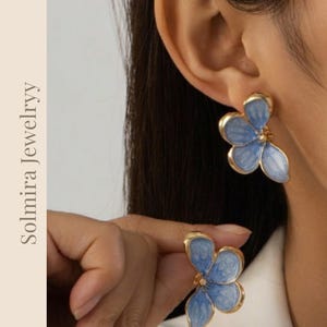 Puede incluir: Pendientes de flores dorados con pétalos azul claro. Los pendientes tienen forma de flor con borde dorado y un pequeño centro dorado. Los pendientes se llevan en la oreja.