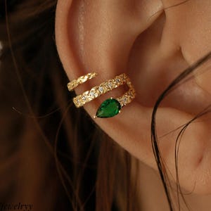 Puede incluir: Un ear cuff dorado con un diseño brillante y una piedra preciosa verde en forma de lágrima. El cuff se envuelve alrededor de la oreja, creando un look elegante. La joya se lleva en la oreja de una persona.