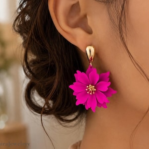 Könnte beinhalten: Nahaufnahme eines goldfarbenen Ohrrings mit einem leuchtend pinkfarbenen Blumenmuster. Die Blume hat mehrere Blütenblätter und eine kleine, goldfarbene Mitte. Der Ohrring ist mit einem goldfarbenen Stecker am Ohr befestigt.