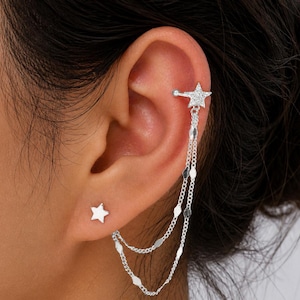May include: A silver ear cuff and stud earring set. The ear cuff features a star design with dangling chains and a star stud earring. The jewelry is designed to be worn on the ear.