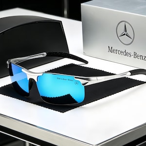 May include: Silver-toned sunglasses with reflective blue lenses and black temple tips. The lenses feature the Mercedes-Benz logo and text. The sunglasses are presented with a silver box displaying the Mercedes-Benz logo and brand name.
