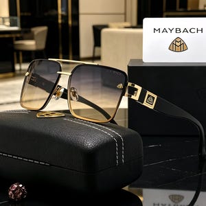 May include: Maybach sunglasses featuring a gold-tone metal frame with a double bridge and square-shaped gradient lenses that transition from dark grey to light amber. The black temples display a gold logo emblem, accompanied by a black textured case.